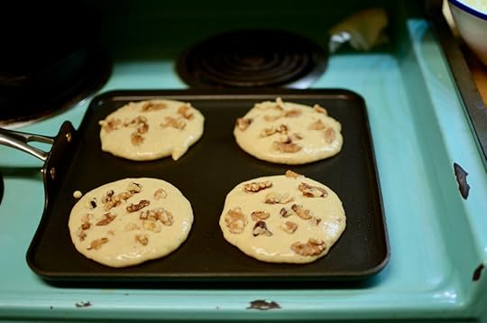 Four pancakes with walnuts tucked into the batter, cooking on a square nonstick griddle. The stove is chipped turquoise enamel.