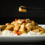 piece of chicken held in black chopsticks over square white plate of chicken stir fry on bed of rice.