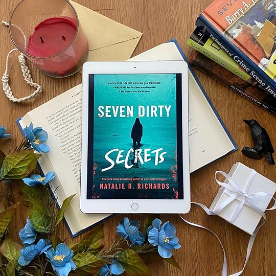 Seven Dirty Secrets book surrounded by blue flowers, crime scene investigation books, and a white box with a white ribbon