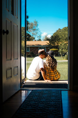A couple is seated on the floor at the threshold of their front door, kissing.