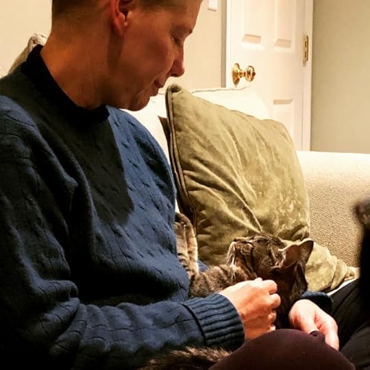 A white woman with short hair sits cross-legged on an ivory sofa with a small tabby cat in her lap gazing at her adoringly