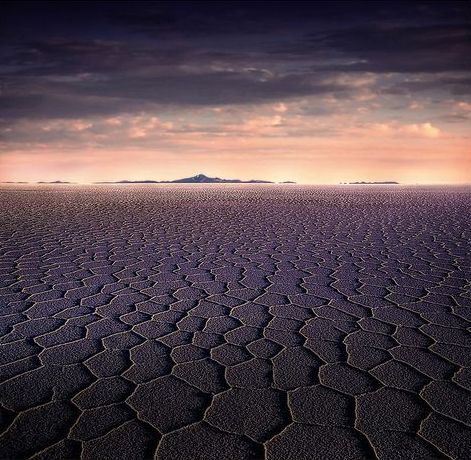 An image of a salt flat in Bolivia, with the salt dried out in a pentagonal pattern, and the shadow gives it a purple tinge. There are mountains at the horizon under a cloud broken sky. Photograph taken by Sergio Pessolano