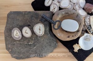 The runes Nauthiz, Kenaz, and Wunjo reversed lay on a large flat stone along with a key and other scattered runes and bones.