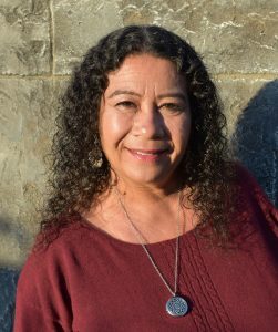 A Latinx woman, Zeena, with black curly hair, and a maroon shirt, smiles at the camera