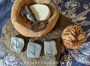The runes Isa, Eihwaz, and Laguz, on a flat brown rock with a carving of a tree and its roots, and a clay bowl holding a nondescript rune and old key.