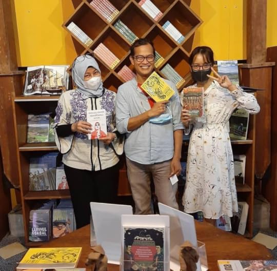 Two women and a man stand in a small bookstore, in front of bookshelves and a brightly painted yellow wall. Each is holding a book and facing the camera, smiling