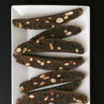 chocolate biscotti with almonds on a white platter on a black table.