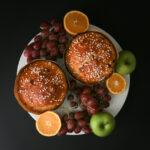 two round loaves of christmas bread on a cake stand surrounded by fruit.