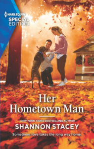 The cover shows a smiling couple playing with a dog against a vibrant autumn background, with fall foliage around them and fallen on the ground. The woman has a long blonde ponytail and is casually dressed in a slouchy pink sweater layered over a white tee, with darker pink jogger pants and white sneakers. She has her hand on the shoulder of a dark-haired man in a long-sleeve gray tee and jeans who is down on one knee petting a large dark brown dog. There’s a large gazebo in the background.
