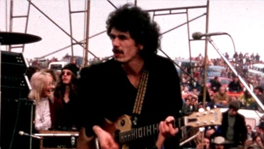 Photo taken from wings of open-air concert of a guitar player, turned away from microphone, with scaffolding and crowds in background under a hazy sky