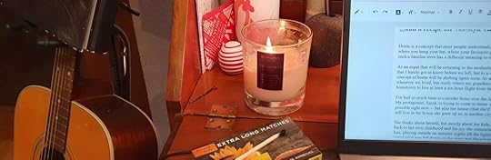 A guitar stands beside a desk that holds a lit candle and a laptop.