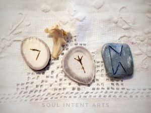 The elder futhark runes Perthro carved into silver clay, Algiz and Kenaz reversed carved into reindeer antler, and a mink vertebra on a white linen background