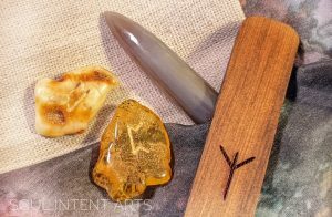 The Elder Futhark runes Algiz in cedar wood, Perthro and Uruz reversed in Baltic amber, with a muddy brown colored Belmnite on a beige and gray background