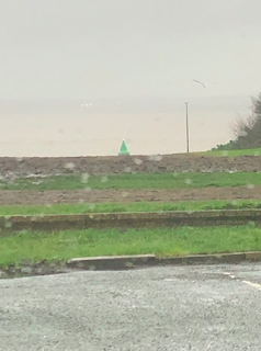 A picture of the Liverpool waterfront on a rainy day, with a grey sky and grey water, and grassy verge leading towards the prom.