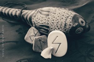 A dark sepia photo of the Elder Futhark runes Sowilo carved into stone, Eihwaz then Raidho reversed carved into clay, with a Mosasaur tooth and owl rattle against a dark background