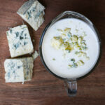 overhead shot of small pitcher of blue cheese dressing on wood board next to chunks of cheese.
