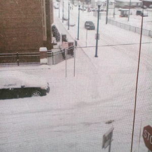 A snowy street scene, taken from a second floor apartment window. Snow covers cars, sidewalk and street. 