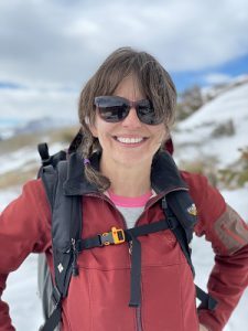 Woman with brown hair, red jacket, sunglasses and backpack with snowy mountains blurred in the background.