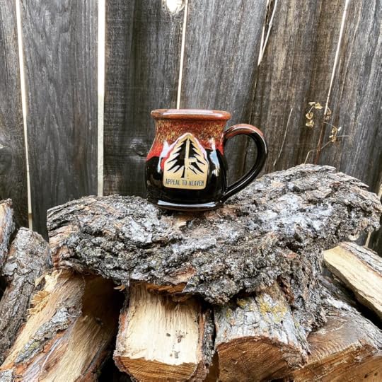 An old-world mug sits on a pile of chopped firewood with a rustic wooden fence in the background. The mug says