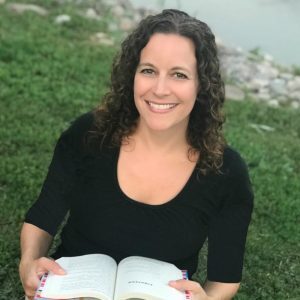 A white woman with dark brown curly hair, Dee Romito, sits in grass, and smiles at the camera while holding a book