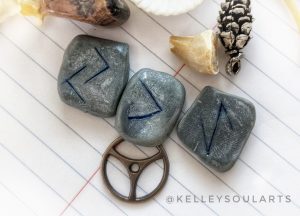 Clay runes Eihwaz, Kenaz reversed, and Jera sit with a metal wheel on white paper with blue lines