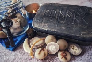 Elder Futhark runes Berkano carved into reindeer antler, then Ingwaz and Laguz painted onto clay runes, lying near a tin with futhark embossed onto it, a beeswax candle, and mink rib, with other runes strewn about