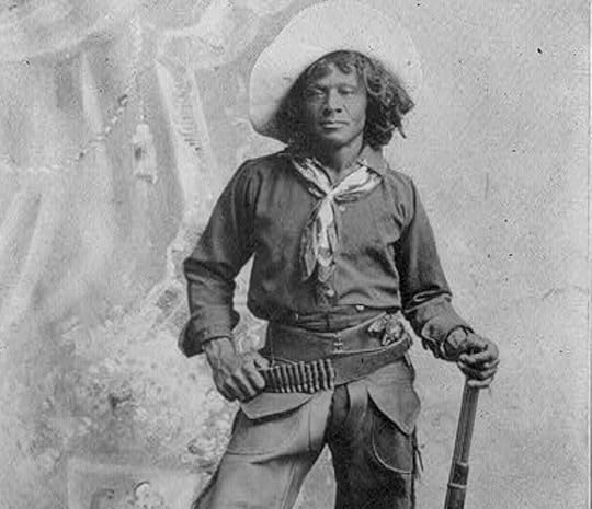 Black and white studio portrait of a cowboy in full work attire, holding a rifle in one hand