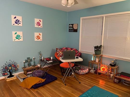 Room with blue walls and hardwood floors. A laptop sits on a small table in front of an orange papasan chair. A small altar and purple meditation cushion are on the left; two large windows with white blinds are on the right.