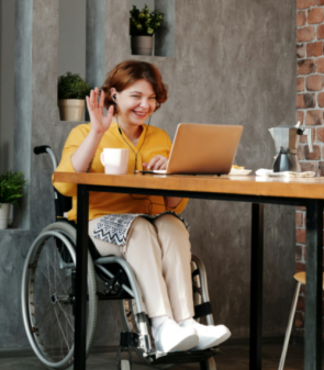 Woman in orange top sitting on black wheelchair. She's looking into her laptop and waving hello.