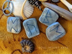 The Elder Futhark runes Berkana, Ehwaz, Uruz, and Perthro reversed on a deerskin background with a fossilized ammonite and other casting bones and shells.