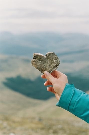 Person standing outdoors holding a heart shaped rock in their hand. The background is a blurry image of mountains and a valley.