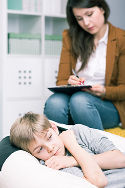 A middle-school age boy lies on a couch in a fetal position while a psychiatrist makes notes.