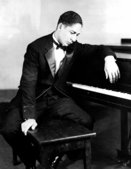 Jelly Roll Morton, in a tuxedo, seated on a piano bench with his left arm draped on top of the piano, looking down at the keys.