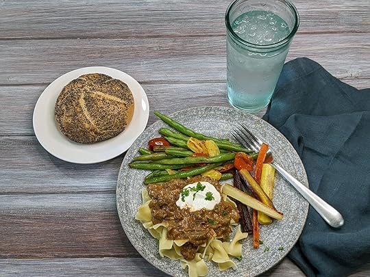 A roll with poppy seeds on a white plate, a plate of ground beef stroganoff over egg noodles with a dollop of sour cream, roasted carrots, and pan seared green beans with multi-colored grape tomatoes, a water glass and a blue napkin