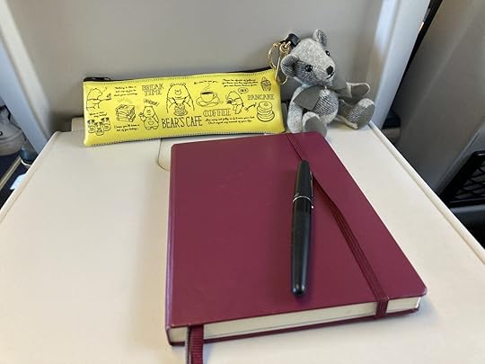 A plastic tray extends back from a train seat. On it, there's a small gray teddy bear, a pencil case that says Bear Cafe on it, and a purple Leuchtermm Journal.
