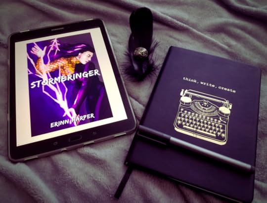 Image ID: a tablet featuring the cover of Erinn Harper’s debut, Stormbringer, situated beside a black journal and pen. The journal’s cover features a typewriter and the words “Think, write, create” in gold.