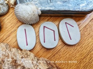 Elder Futhark runes Lagyz, Uruz, and Isa carved into white ceramic tiles with a sea shell, on a light wooden background, with a rough chunk of wood beneath them.