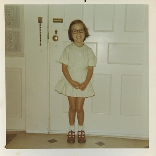 Ayun Halliday, age 5, posing before her family's front door in her pale green St. Richards School uniform, white socks, red English sandals, and teeny tiny glasses.