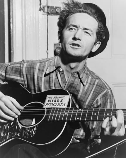 Black and white portrait of Woody Guthrie, seated, playing a guitar