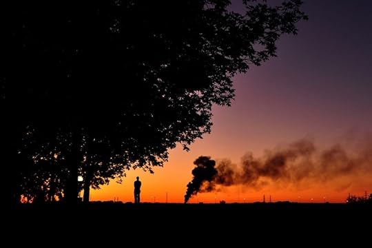 Image: a lone person stands watching distant smoke rising, silhouetted against a colorful sunrise.