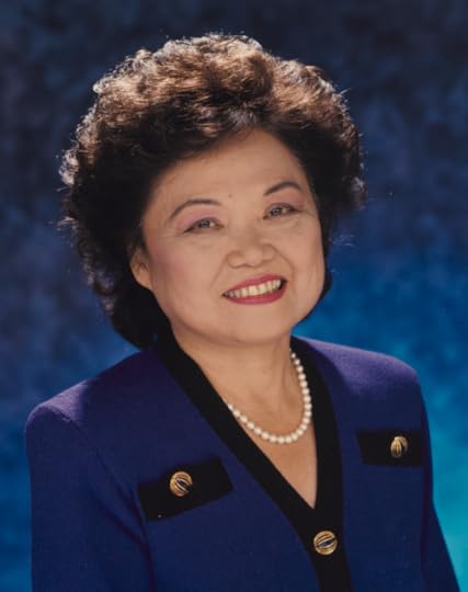 Head and shoulders color portrait of Patsy Mink, wearing a blue blazer and a strand of pearls, smiling, against a blue blackground