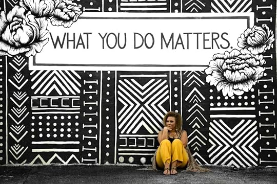 Image: a woman sitting against a wall on which is painted a black and white mural and the words 