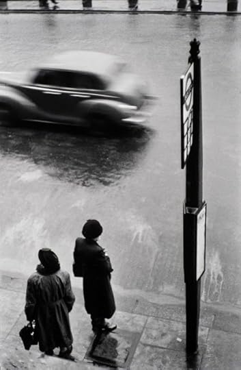 A black and white photo of two women in long coats and hats standing at a bus stop in London, in 1952, with a blurred car on the road passing by. Taken by Elliot Erwitt