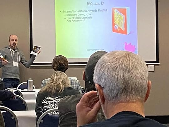 Ryan R. Campbell holds his book AND AMPERSAND in one hand while standing alongside a projected screen that displays his book SCAMBAIT
