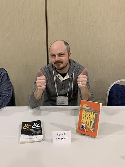 Ryan R. Campbell sits at a table with copies of his books SCAMBAIT and AND AMPERSAND displayed in front of him.