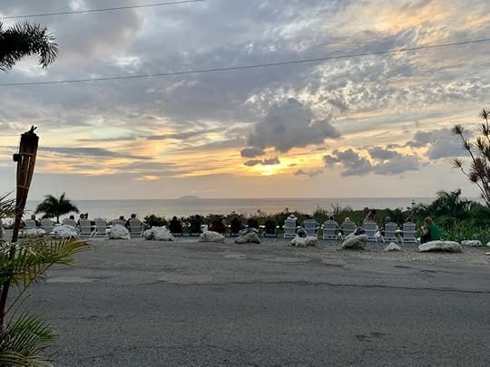 A row of chairs overlook a drop-off to the sea, where the sun sets on the horizon.
