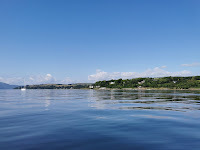 a sunny day looking onto the shore of Loch Long. The water is pale blue and a rolling hill is in the distance.