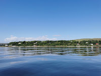a sunny day looking onto the shore of Loch Long. The water is pale blue and a rolling hill is in the distance.