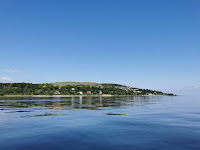 A sunny day looking onto the shore of Loch Long. The water is pale blue and a rolling hill is in the distance.