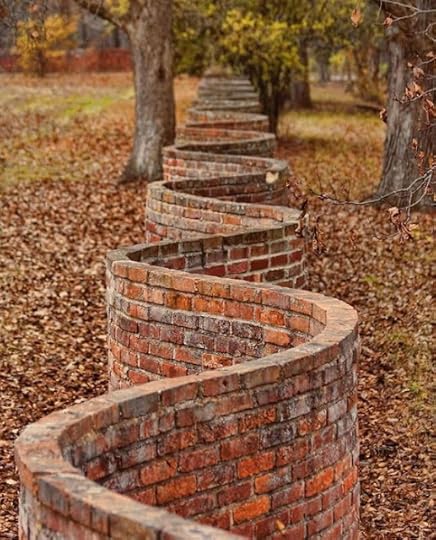 An arial view of a terracotta-coloured, single brick wall, built so that it goes in and out, like a squiggle. It's called a Crinkle Crankle wall. It is across a wooded landscape in autumn.